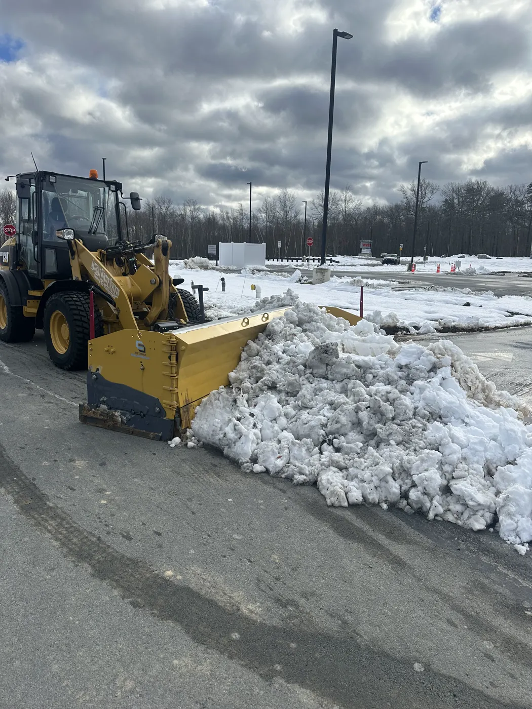 Yellow wheel loader pushing snow into a pile at the edge of a lot