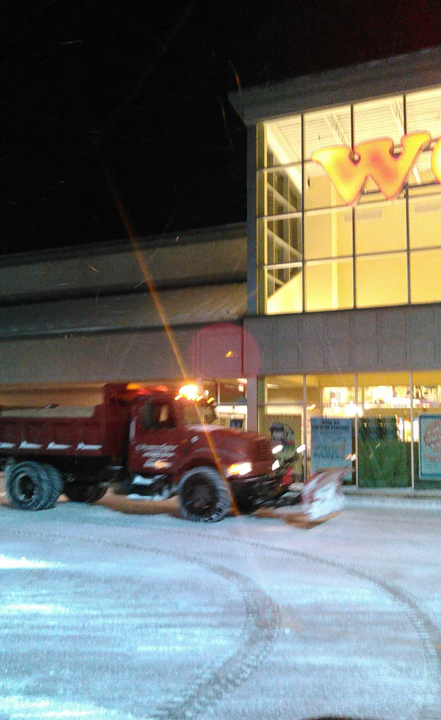 Red dump truck with plow clearing the lot in front of a lit retail store at night